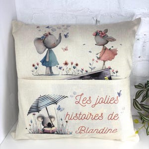 May include: A cushion with a pocket, decorated with illustrations of an elephant and a deer in dresses, amidst flowers and butterflies. The lower part displays a koala under an umbrella, with the text "Les jolies histoires de Blandine."