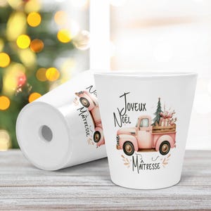 Könnte beinhalten: Zwei weiße Keramikbecher mit einer Illustration eines rosa Vintage-Pickups, der einen Weihnachtsbaum und Geschenke geladen hat. Der Text Joyeux Noël Maîtresse ist in schwarzer Schrift zu sehen. Das Design enthält florale Akzente.
