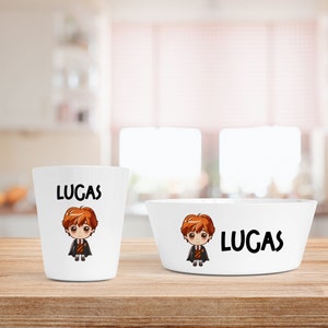 May include: White ceramic bowl and cup with a cartoon character and the name "Lucas" printed on them. The bowl and cup are on a wooden surface.