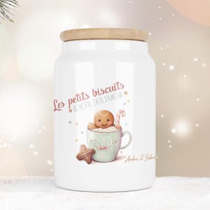 Peut inclure: Un bocal en céramique blanche avec un couvercle en bois, orné d'un dessin de bonhomme en pain d'épices dans une tasse vert clair, d'un sucre d'orge et d'un biscuit en étoile. Le texte indique Les petits biscuits de notre tatie d'amour.