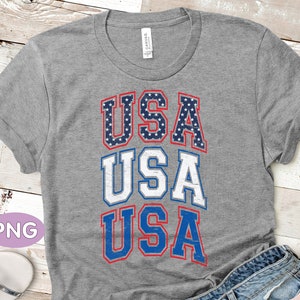 May include: Gray t-shirt with a distressed graphic of the word "USA" repeated three times. The letters are outlined in blue and red, and the "A" in each word is decorated with stars.