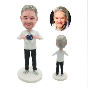 Op de afbeelding: Een gepersonaliseerd bobblehead-beeldje van een persoon met grijs haar, die een wit shirt draagt met een blauw-wit logo op de borst. Het beeldje houdt het shirt open om het logo te laten zien. Het beeldje staat op een witte voet.
