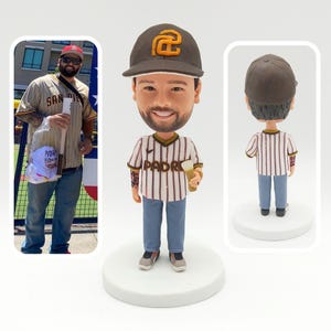 Op de afbeelding: Een gepersonaliseerd bobblehead-beeldje van een persoon die een bruine en gouden baseballpet draagt met de letter "G" erop, een wit en bruin gestreept shirt met het woord "PADRE" erop, een blauwe jeans en een klein kopje vasthoudt. Het beeldje staat op een witte voet.