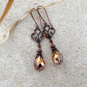 May include: A pair of dangle earrings featuring antique copper-toned metal filigree scrollwork tops and faceted, teardrop-shaped amber-colored glass beads suspended from the bottom. The earrings have curved ear wires for secure placement.