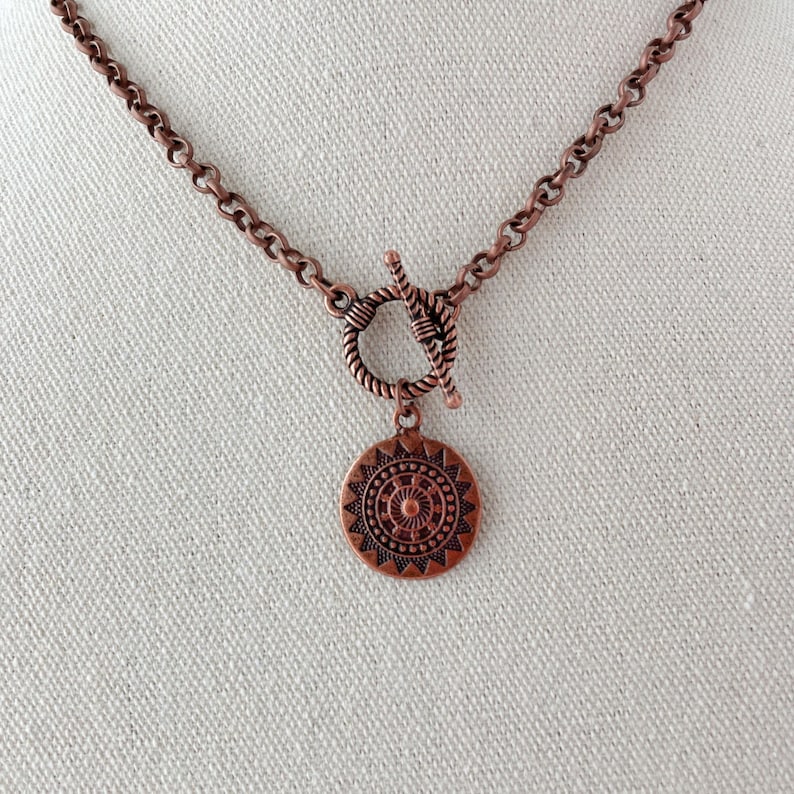 May include: A copper-toned necklace featuring a round, textured pendant with a radial, sun-like pattern. The pendant hangs from a circular toggle clasp, which is connected to a thick, round-link chain, all finished in a matching copper metal.