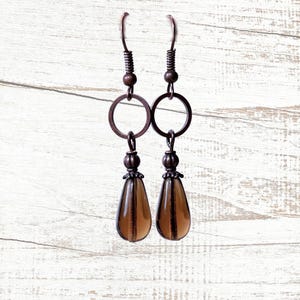 May include: A pair of copper-colored earrings. Each earring features a teardrop-shaped, amber-colored glass bead suspended from a copper ring. The earrings have a decorative bead and a coiled wire at the top, with a hook for the ear.
