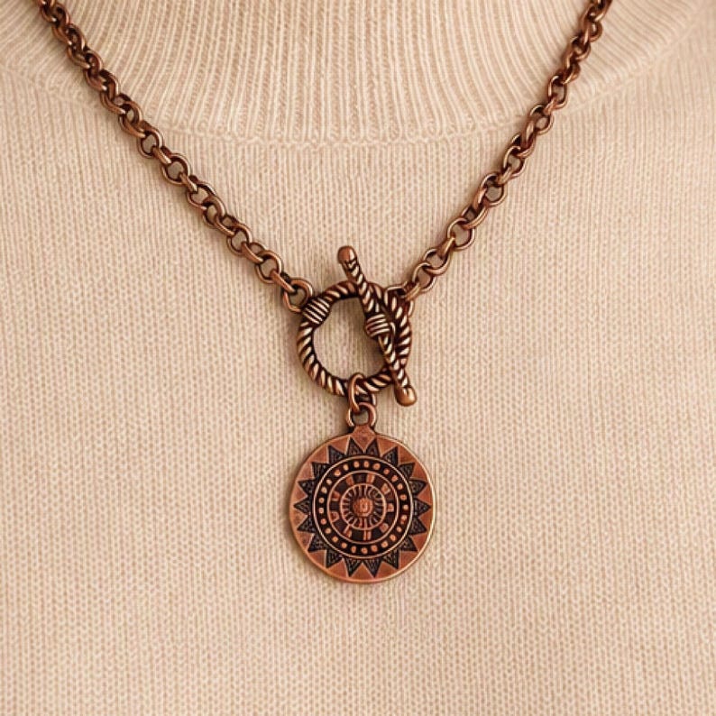 May include: A copper-toned necklace featuring a round pendant with a sunburst pattern and geometric engravings. The pendant hangs from a thick cable chain secured by a rope-textured toggle clasp, creating a cohesive metallic look throughout.