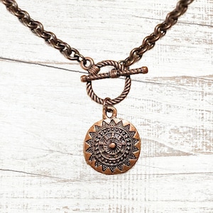 May include: A copper-colored necklace with a circular pendant featuring a sunburst design. The pendant hangs from a textured chain with a toggle clasp. The necklace is displayed against a white wooden background.