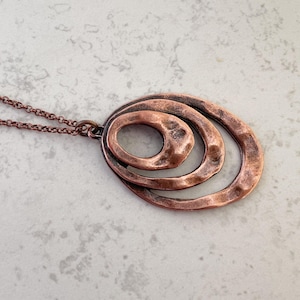 May include: Copper-colored pendant necklace featuring an abstract design of nested ovals. The hammered texture adds visual interest to the pendant, which hangs from a delicate chain. A unique piece of jewelry.