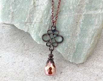Könnte beinhalten: Eine kupferfarbene Halskette mit einer feinen Kette und einem kunstvollen, durchbrochenen Metallanhänger in einem geschwungenen, symmetrischen Design. Eine facettierte, tropfenförmige Perle in Kupferoptik hängt an der Basis des Anhängers.