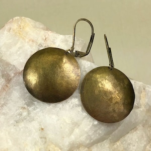 May include: A pair of antique brass-toned disc earrings. Each earring features a large, hammered, circular design with a lever-back closure. The earrings have a vintage, tarnished appearance, suggesting age and wear. The earrings are displayed on a white, textured surface.