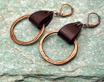 May include: A pair of handcrafted earrings featuring large, circular copper hoops. Each earring has a dark brown leather accent at the top, connected to a copper-colored lever-back closure. The earrings rest on a textured, light green surface.