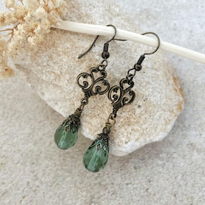 May include: A pair of dangle earrings featuring antique bronze-toned metal filigree scrollwork connectors attached to faceted, teardrop-shaped sage green glass beads. The earrings hang from simple bronze-toned metal ear wires.