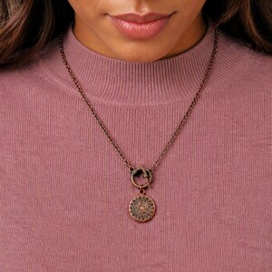 May include: A copper-toned metal necklace featuring a circular pendant with a sunburst or compass-like engraved design. The pendant hangs from a matching chain that connects to a circular toggle clasp mechanism positioned at the center front.