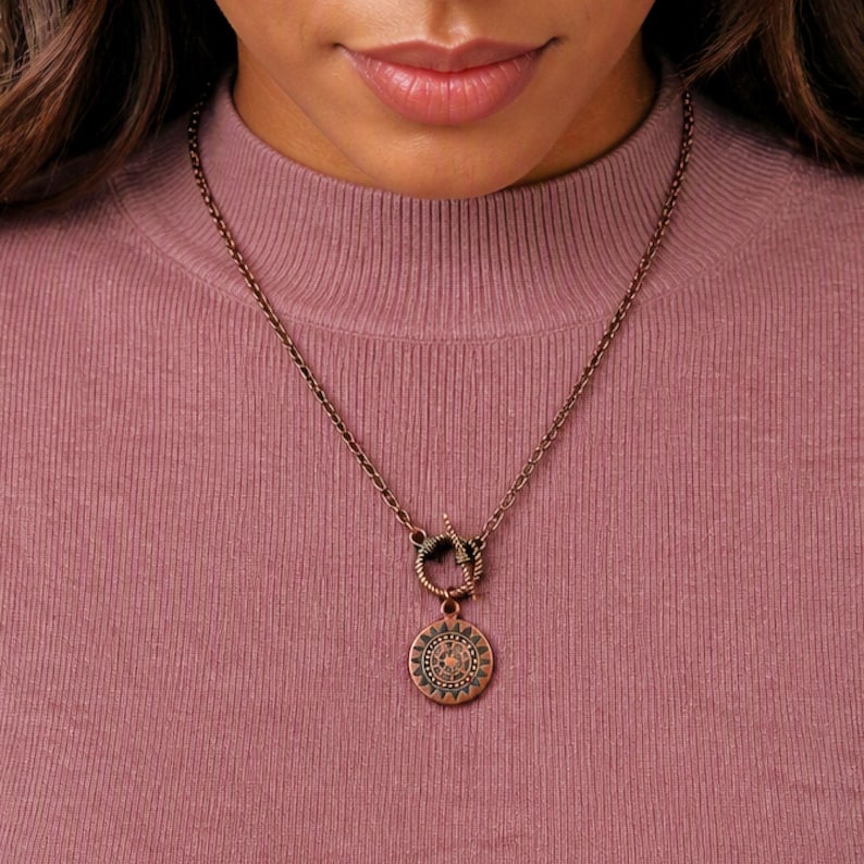 May include: A copper-toned metal necklace featuring a circular pendant with a sunburst or compass-like engraved design. The pendant hangs from a matching chain that connects to a circular toggle clasp mechanism positioned at the center front.