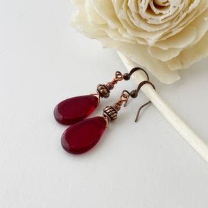 May include: A pair of drop earrings featuring deep red, teardrop-shaped glass beads suspended from copper-toned wire hooks. Each earring includes a decorative, ribbed copper-colored metal bead cap positioned directly above the smooth red glass drop.