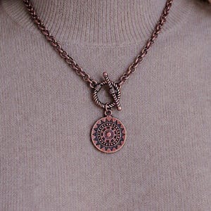 May include: A copper-toned metal chain necklace featuring a toggle clasp with a circular pendant. The pendant displays a detailed, symmetrical sunburst or mandala-style geometric pattern with radiating lines and small circular accents throughout.