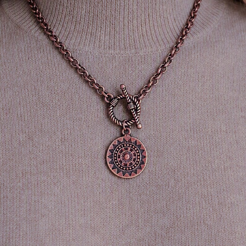 May include: A copper-toned metal chain necklace featuring a toggle clasp with a circular pendant. The pendant displays a detailed, symmetrical sunburst or mandala-style geometric pattern with radiating lines and small circular accents throughout.