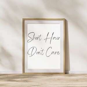 Puede incluir: Una impresión enmarcada con el texto "Short Hair Don't Care" en una fuente cursiva. El marco es de madera clara y el fondo es blanco.