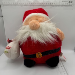 Puede incluir: Un juguete de peluche de Papá Noel que lleva un traje rojo con barba blanca y cinturón negro. Lleva una bolsa blanca con la palabra "Navidad" en ella.
