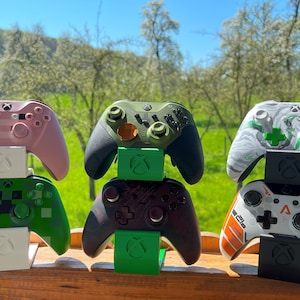 Puede incluir: Cuatro mandos de Xbox de diferentes colores se muestran en soportes individuales. Los mandos están apilados en pares, con un mando rosa encima de un mando verde, un mando verde encima de un mando negro y un mando blanco encima de un mando blanco. Los mandos están todos en un entorno natural con un campo verde y árboles en el fondo.