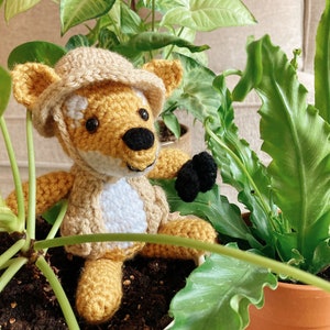 Może przedstawiać: Szydełkowy lis w beżowym kapeluszu siedzi w doniczce z zielonymi roślinami. Lis jest żółty i biały z czarnymi oczami i czarnym nosem.