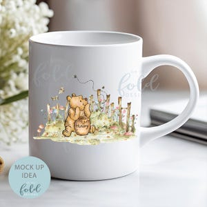 May include: White ceramic mug featuring an illustration of Winnie the Pooh. Pooh is holding a honey pot and standing in a field of flowers with a fence behind him. The mug has the text "on the fold designs" on the side.