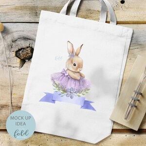 Peut inclure: Sac fourre-tout blanc avec une illustration aquarelle de lapin en tutu et couronne de fleurs violettes. Le lapin tient une fleur violette dans sa patte. Le sac fourre-tout a un ruban bleu avec le texte "fold" dessus.