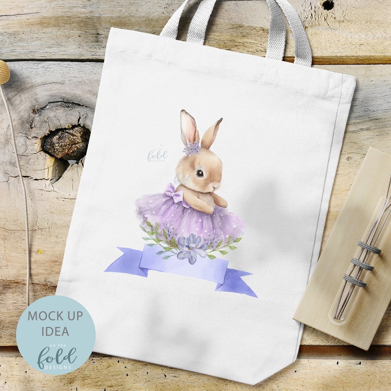 Peut inclure: Sac fourre-tout blanc avec une illustration aquarelle de lapin en tutu et couronne de fleurs violettes. Le lapin tient une fleur violette dans sa patte. Le sac fourre-tout a un ruban bleu avec le texte "fold" dessus.