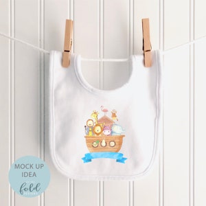 May include: White baby bib with a colorful illustration of Noah's Ark, featuring various animals and a blue banner. The bib is hanging on a clothesline with wooden clothespins. The words "MOCK UP IDEA" and "on the fold designs" are printed on a blue circle.