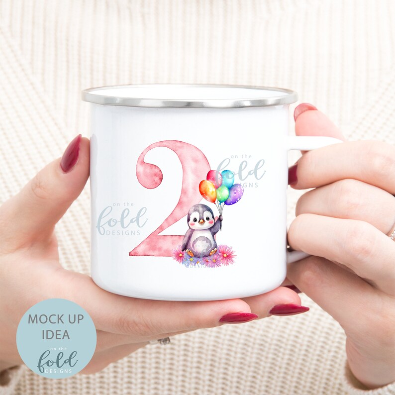 May include: A white enamel mug with a pink number two and a penguin holding colorful balloons. The mug has the text "on the fold designs" on it.