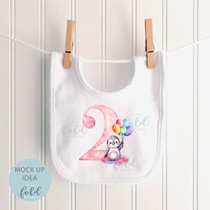 May include: White baby bib with a pink number two and a penguin holding colorful balloons. The text "On the Fold Designs" is printed on the bib.