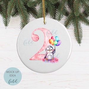 May include: A pink and white ceramic ornament with a penguin holding colorful balloons. The number "2" is in the background with the text "on the fold designs" on the ornament.