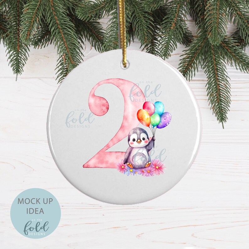 May include: A pink and white ceramic ornament with a penguin holding colorful balloons. The number "2" is in the background with the text "on the fold designs" on the ornament.