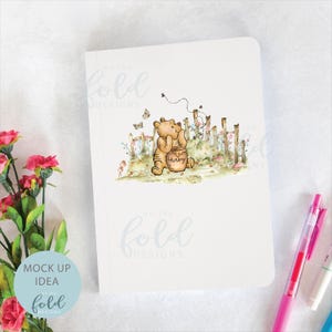 May include: A white notebook featuring a watercolour illustration of Winnie the Pooh holding a honey pot. The illustration is surrounded by flowers and a fence. The text "On the Fold Designs" is printed in light blue at the top and bottom of the notebook.