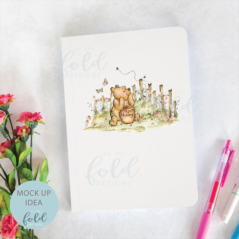 May include: A white notebook featuring a watercolour illustration of Winnie the Pooh holding a honey pot. The illustration is surrounded by flowers and a fence. The text "On the Fold Designs" is printed in light blue at the top and bottom of the notebook.