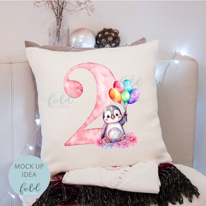 May include: A white pillow with a pink number two and a penguin holding colorful balloons. The text "On the Fold Designs" is in the bottom left corner of the pillow.
