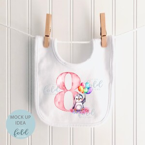 May include: White bib with a pink number eight and a penguin holding rainbow balloons. The text "On the Fold Designs" is on the bib.