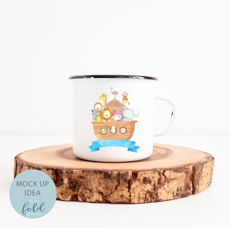 May include: White enamel mug with a black rim, featuring a colorful illustration of Noah's Ark with various animals. A blue banner is below the ark. The mug is on a wooden slice. Text on the bottom left says "MOCK UP IDEA on the fold DESIGNS".