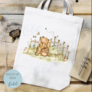 May include: White tote bag featuring a watercolour illustration of Winnie the Pooh holding a honey pot. The illustration is surrounded by flowers and a fence. The text "Hunny" is on the honey pot.