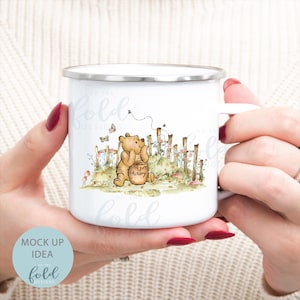 May include: White enamel mug featuring an illustration of Winnie the Pooh. Pooh is holding a honey pot and looking at a bee flying towards him. The illustration is surrounded by flowers and a fence.