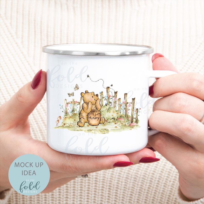 May include: White enamel mug featuring an illustration of Winnie the Pooh. Pooh is holding a honey pot and looking at a bee flying towards him. The illustration is surrounded by flowers and a fence.
