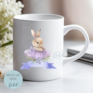 Peut inclure: Mug en c&eacute;ramique blanche avec une illustration aquarelle d'un lapin brun portant un tutu violet et un ruban bleu. Le texte "on the fold designs" est imprim&eacute; sur la tasse.