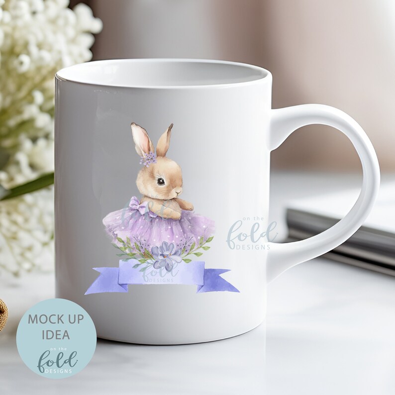 Peut inclure: Mug en c&eacute;ramique blanche avec une illustration aquarelle d'un lapin brun portant un tutu violet et un ruban bleu. Le texte "on the fold designs" est imprim&eacute; sur la tasse.