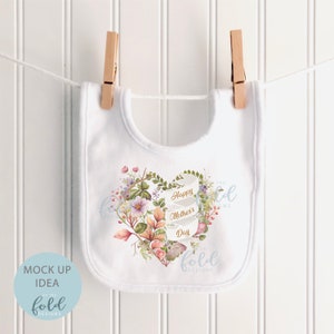 May include: White baby bib with a floral heart design and the text "Happy Mother's Day" in gold lettering. The design includes a heart shape made of leaves and flowers in shades of pink, purple, and green. The text "Like the Fold Designs" is also included in the design.