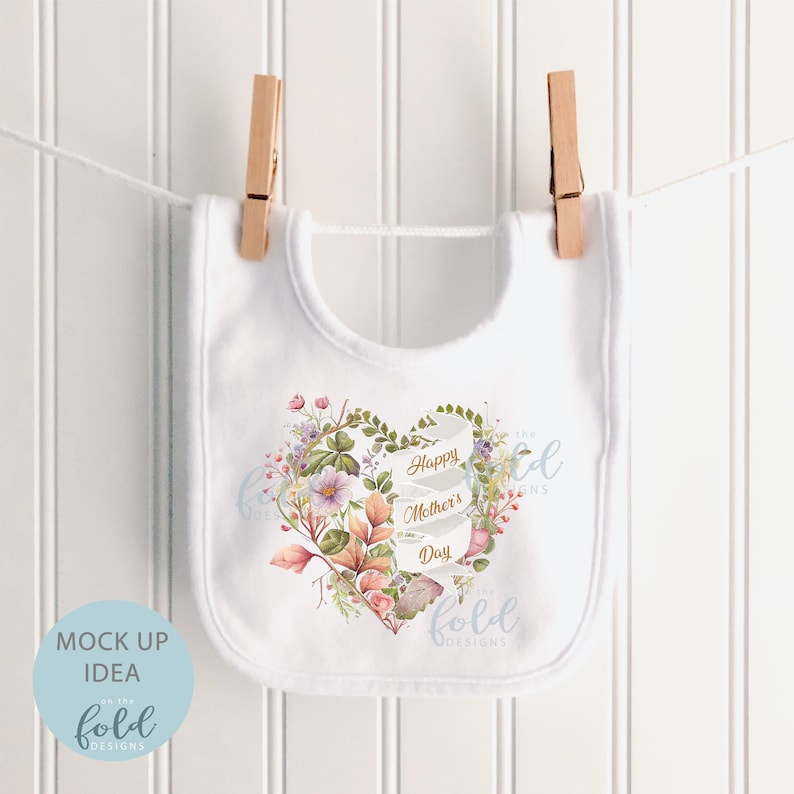 May include: White baby bib with a floral heart design and the text "Happy Mother's Day" in gold lettering. The design includes a heart shape made of leaves and flowers in shades of pink, purple, and green. The text "Like the Fold Designs" is also included in the design.