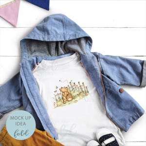 May include: White t-shirt featuring a cartoon illustration of Winnie the Pooh holding a pot of honey. The image is set against a backdrop of a fence and flowers. The t-shirt is displayed on a blue denim jacket and brown trousers.