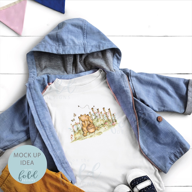 May include: White t-shirt featuring a cartoon illustration of Winnie the Pooh holding a pot of honey. The image is set against a backdrop of a fence and flowers. The t-shirt is displayed on a blue denim jacket and brown trousers.