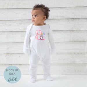 May include: White long-sleeve baby onesie with a pink number eight and a penguin holding colorful balloons. The onesie has the text "Hola" on the sleeve.