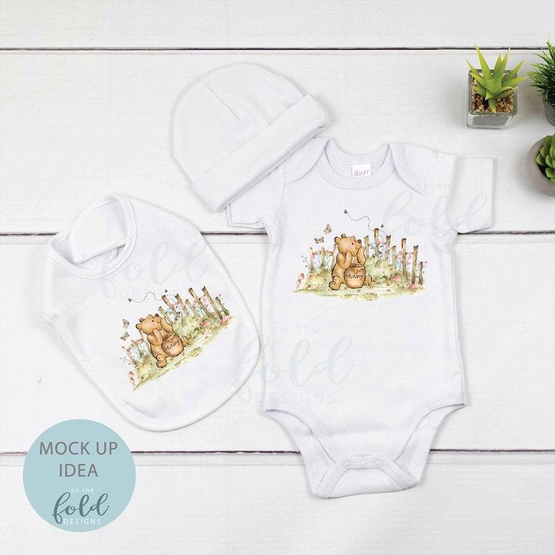 May include: A white baby bodysuit, bib and hat set featuring a Winnie the Pooh illustration. The illustration shows Winnie the Pooh holding a honey pot in a woodland setting.